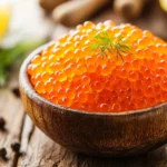 A wooden bowl filled with fresh, vibrant orange salmon roe, garnished with a sprig of dill, surrounded by rustic table decor.