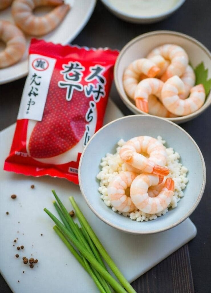 ngredients for a Boston Roll Sushi recipe, featuring fresh cooked shrimp placed on a bed of rice in a small ceramic bowl, with another bowl of shrimp nearby. A red packet of tobiko (fish roe), fresh green scallions, and scattered peppercorns are displayed on a white cutting board.
