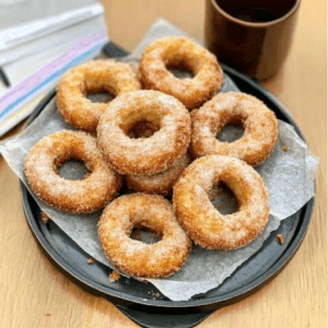 A plate of crispy, Crispy Chicken and Potato doughnuts coated with sugar, served on parchment paper with a cup of coffee in the background.