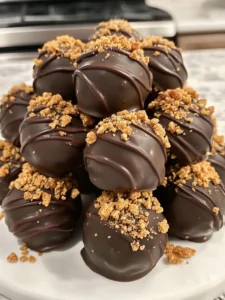 Close-up of Butterfinger Balls stacked on a white plate, coated in rich chocolate and topped with crushed Butterfinger candy.