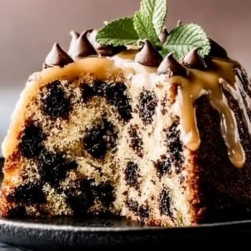 A moist slice of Chocolate Chip Banana Cake Bliss topped with caramel glaze, chocolate chips, and mint leaves, displayed on a black plate.