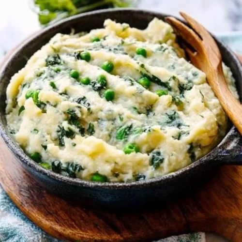 A rustic black bowl filled with Creamy Dreamy Colcannon Mash-Up mashed potatoes blended with greens and green peas, served on a wooden board with a wooden fork.