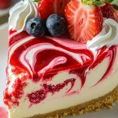 A slice of Dreamy Strawberry Swirl Cheesecake with vibrant red swirls, fresh strawberries, blueberries, and whipped cream on top