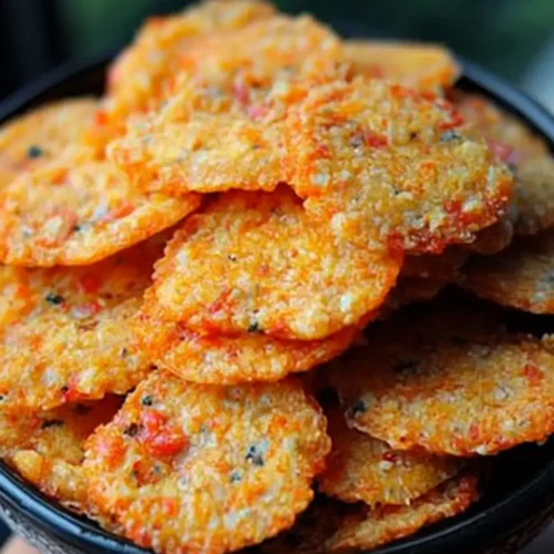 A bowl filled with thin, crispy pimento cheese crisps, golden-orange in color with flecks of red pepper and herbs.