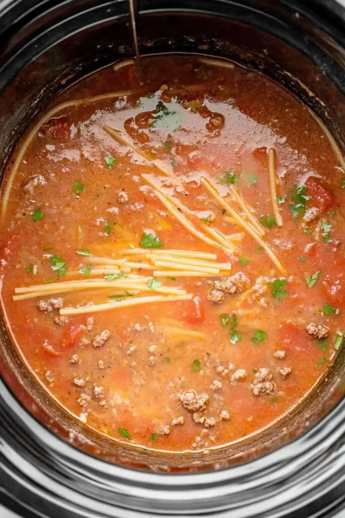 Crockpot taco spaghetti with cream cheese Close-up view of Crockpot taco spaghetti with cream cheese in the early cooking stage, showing uncooked spaghetti noodles in a rich, orange-red tomato sauce with ground beef, flecks of green herbs, and the beginning stages of cream cheese melting into the mixture.