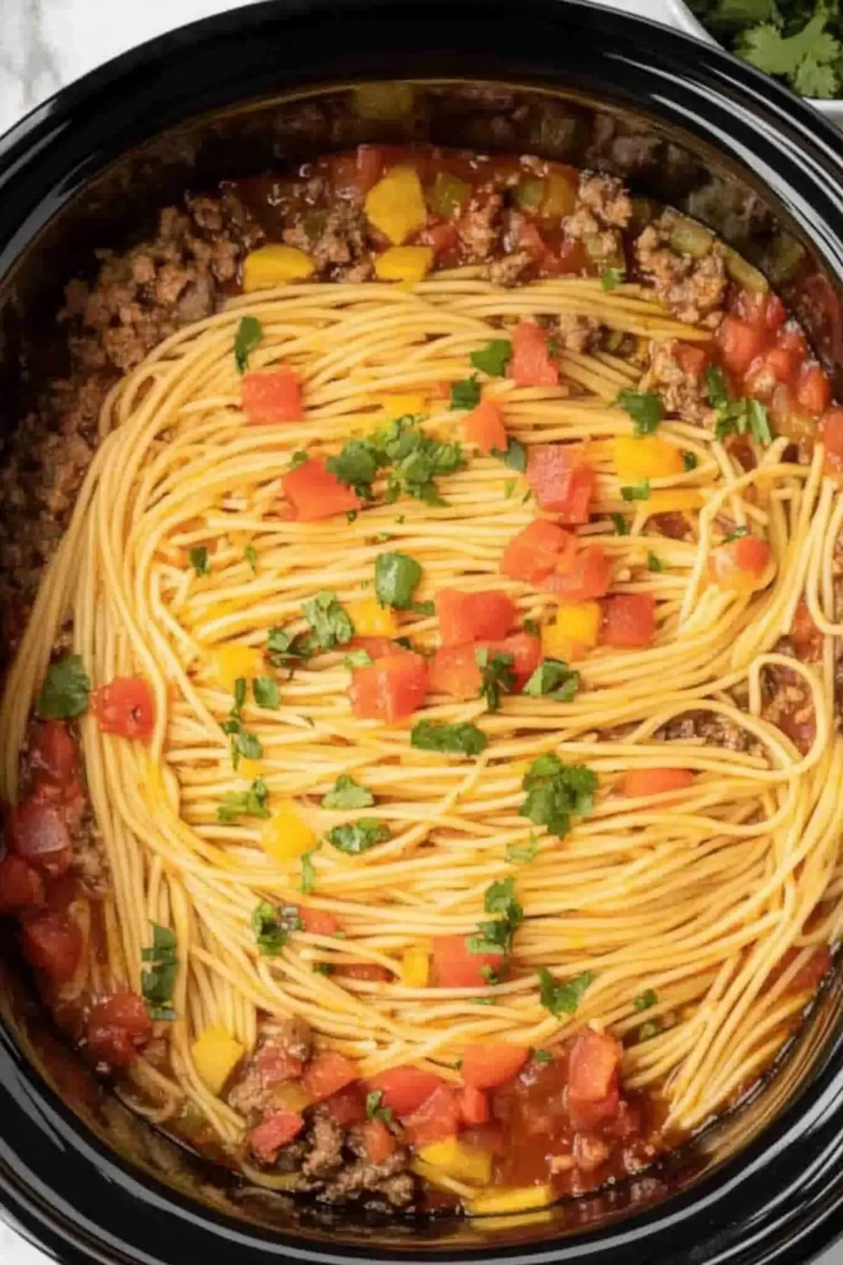 Crockpot taco spaghetti with ground beef Overhead view of Crockpot taco spaghetti with ground beef almost fully cooked, featuring cooked spaghetti noodles on top with diced red and yellow bell peppers, fresh cilantro, and seasoned ground beef visible around the edges in a savory tomato sauce.