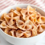 A white bowl filled with golden-brown bow tie Easy Air Fryer Churro Pasta Chips Recipe coated in cinnamon sugar, resembling churros, served on a marble surface with a red and white striped cloth in the background.