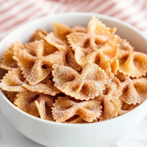 A white bowl filled with golden-brown bow tie Easy Air Fryer Churro Pasta Chips Recipe coated in cinnamon sugar, resembling churros, served on a marble surface with a red and white striped cloth in the background.