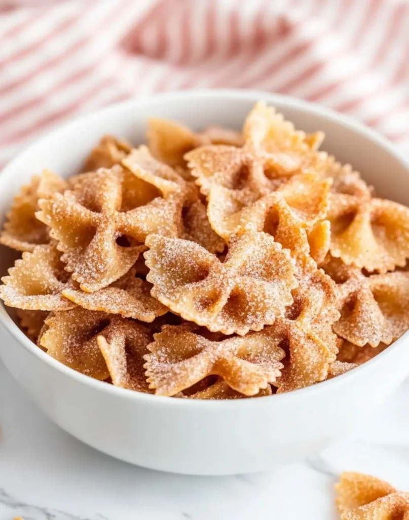 A white bowl filled with golden-brown bow tie Easy Air Fryer Churro Pasta Chips Recipe coated in cinnamon sugar, resembling churros, served on a marble surface with a red and white striped cloth in the background.