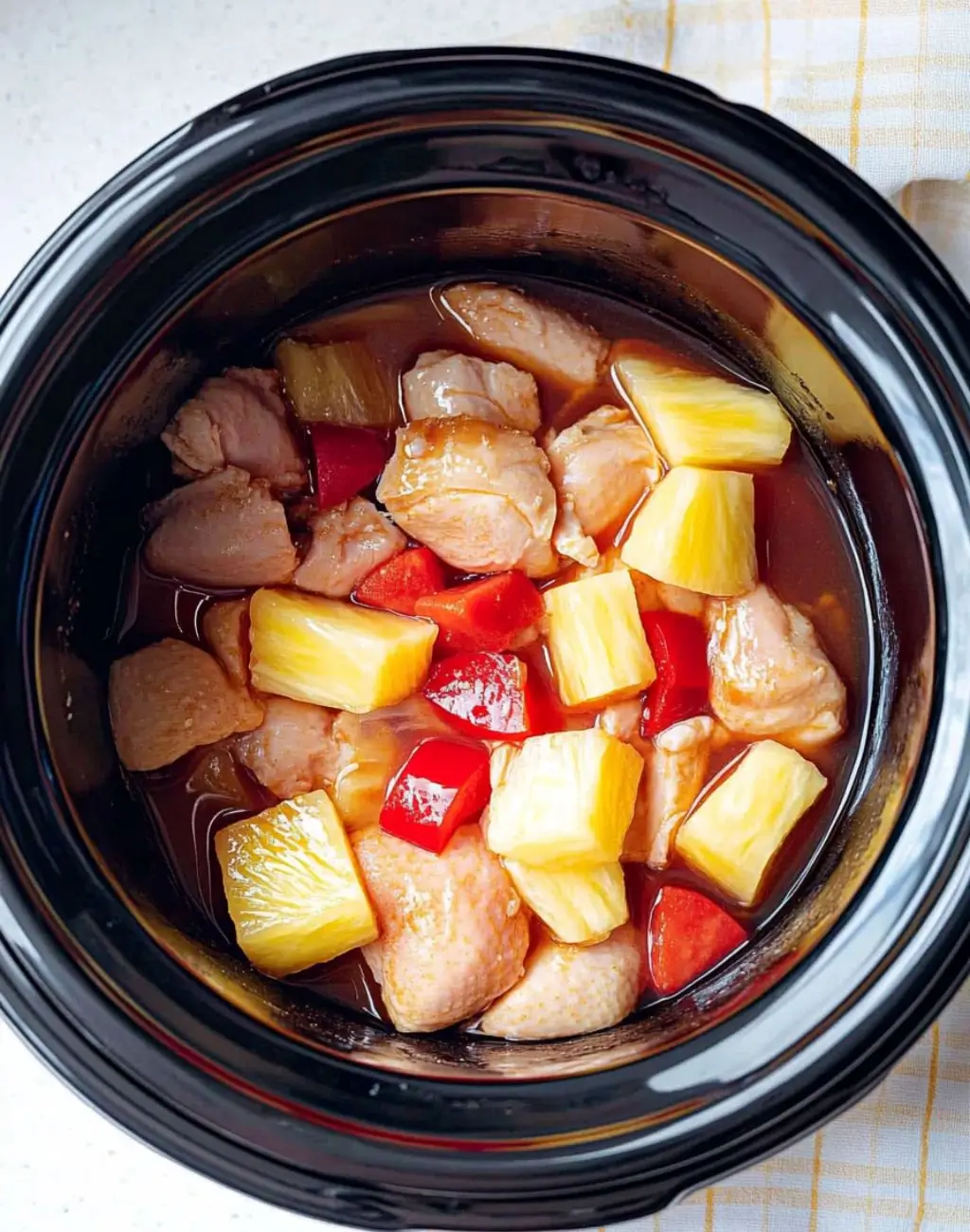 Hawaiian Crockpot Chicken Overhead view of raw chicken pieces, fresh pineapple chunks, and red bell peppers in a black slow cooker with Hawaiian sauce, ready to be cooked.