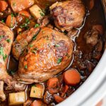 Close-up view of Slow Cooker Chicken Merlot with golden-brown seared chicken thighs, mushrooms, carrots, and potatoes in rich dark wine sauce, garnished with fresh parsley in white Dutch oven