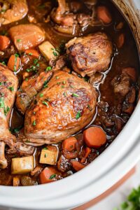Close-up view of Slow Cooker Chicken Merlot with golden-brown seared chicken thighs, mushrooms, carrots, and potatoes in rich dark wine sauce, garnished with fresh parsley in white Dutch oven