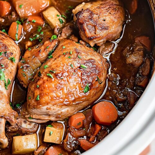Close-up view of Slow Cooker Chicken Merlot with golden-brown seared chicken thighs, mushrooms, carrots, and potatoes in rich dark wine sauce, garnished with fresh parsley in white Dutch oven