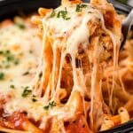 A forkful of Slow Cooker Chicken Parmesan Pasta being lifted from a black bowl, with melted mozzarella stretching in long, gooey strings. The pasta appears to be penne in a rich tomato sauce, garnished with fresh herbs.