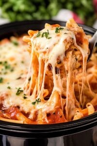 A forkful of Slow Cooker Chicken Parmesan Pasta being lifted from a black bowl, with melted mozzarella stretching in long, gooey strings. The pasta appears to be penne in a rich tomato sauce, garnished with fresh herbs.
