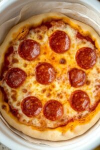 A homemade pepperoni Slow Cooker Pizza, showing golden melted cheese, perfectly arranged pepperoni slices, and a golden-brown crust, all contained within a white slow cooker lined with parchment paper.
