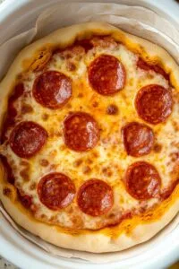 A homemade pepperoni Slow Cooker Pizza, showing golden melted cheese, perfectly arranged pepperoni slices, and a golden-brown crust, all contained within a white slow cooker lined with parchment paper.