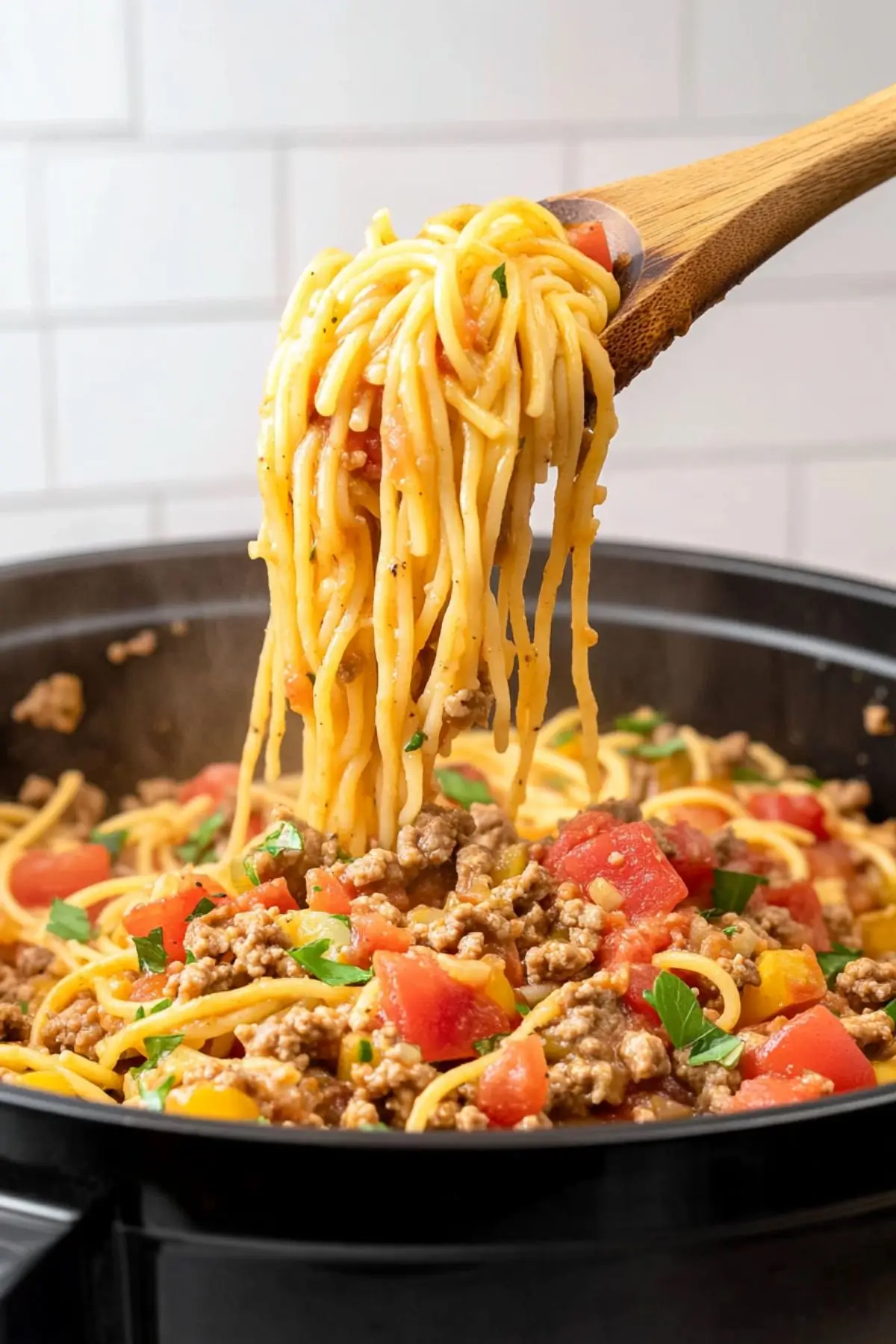 Taco spaghetti recipe Close-up of taco spaghetti being lifted from a black skillet with a wooden spoon, showing spaghetti noodles coated in cheese sauce with seasoned ground beef, diced tomatoes, and fresh herbs visible throughout the dish.