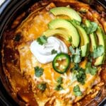 Slow cooker chicken enchilada casserole topped with melted cheese, fresh avocado slices, sour cream, jalapeño, and cilantro in a black crockpot.