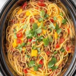 Overhead view of crockpot taco spaghetti with spaghetti noodles, seasoned ground beef, diced red and yellow bell peppers, all garnished with fresh cilantro. Bowls of shredded cheese and chopped herbs visible around the edges.