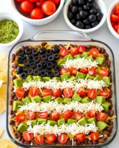 American Flag Layered Taco Dip decorated with black olives, diced tomatoes, shredded cheese, and avocado to resemble the U.S. flag in a glass dish.