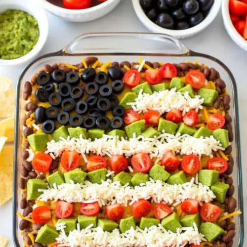 American Flag Layered Taco Dip decorated with black olives, diced tomatoes, shredded cheese, and avocado to resemble the U.S. flag in a glass dish.