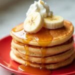 Fluffy Cottage Cheese Pancakes stacked on a red plate, topped with banana slices, whipped cream, and drizzled maple syrup.