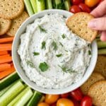 Cottage cheese dip recipes with fresh herbs served in a white bowl, surrounded by crackers, cherry tomatoes, cucumber, and carrot sticks.